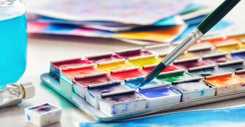 Set of watercolor paints, paintbrush, glass of water and paper sheets of watercolor paintings on background. Selective focus.